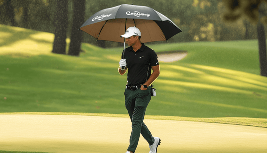 A golfer crosses the green while holding a golf umbrella to protect from the sun.