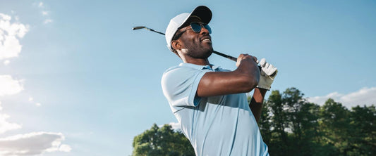 Golfer in a blue shirt and sunglasses holding a club after a swing, with a clear sky and trees in the background.